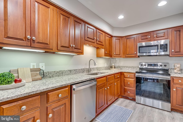 kitchen with appliances with stainless steel finishes, a sink, brown cabinetry, light stone counters, and light wood-style flooring
