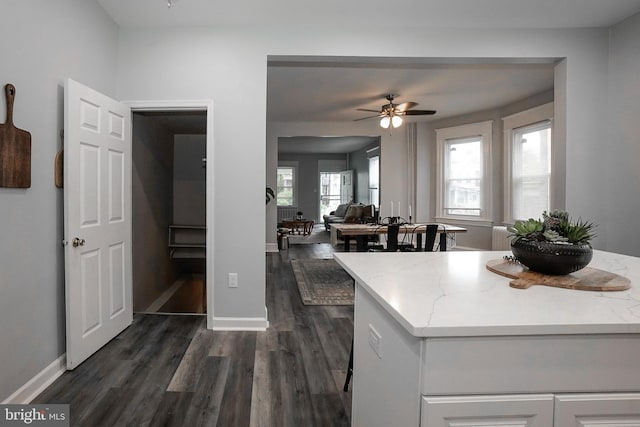kitchen with white cabinets, light stone countertops, a ceiling fan, baseboards, and dark wood-type flooring
