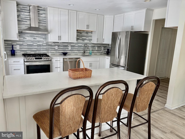 kitchen with light wood-style floors, a kitchen breakfast bar, wall chimney range hood, a sink, and appliances with stainless steel finishes