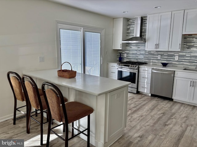 kitchen featuring a kitchen bar, light wood-type flooring, wall chimney exhaust hood, appliances with stainless steel finishes, and backsplash