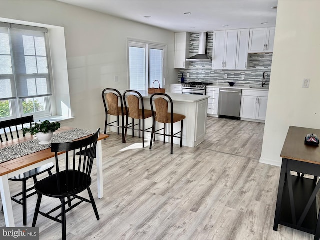 kitchen featuring wall chimney range hood, appliances with stainless steel finishes, light wood-style floors, a kitchen breakfast bar, and plenty of natural light
