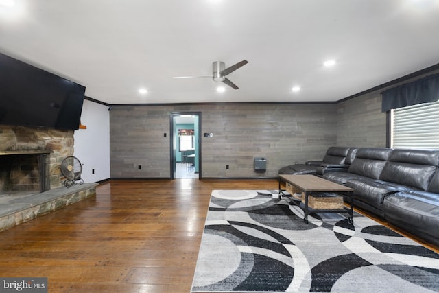 living room with crown molding, wood-type flooring, a stone fireplace, a ceiling fan, and recessed lighting