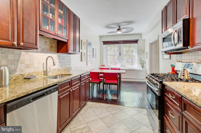 kitchen featuring light stone counters, a sink, stainless steel appliances, a ceiling fan, and reddish brown cabinets