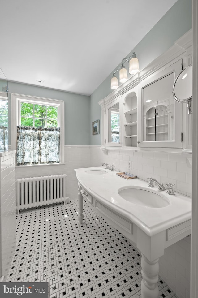 bathroom with tile patterned flooring, wainscoting, dual sinks, tile walls, and radiator heating unit