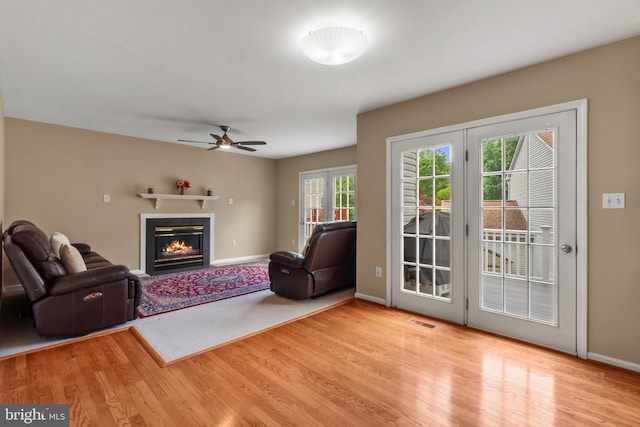 living area featuring wood finished floors, a glass covered fireplace, baseboards, and a ceiling fan