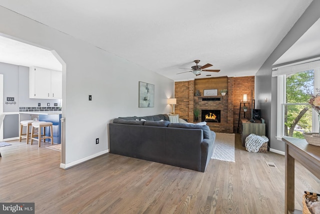 living room with ceiling fan, light wood finished floors, a brick fireplace, arched walkways, and baseboards