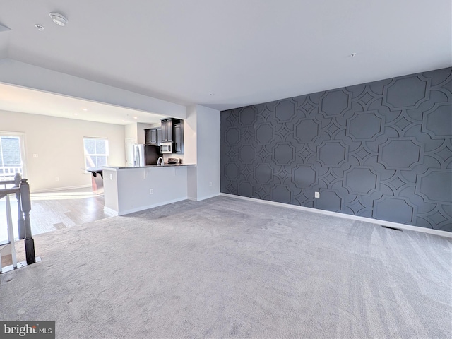 unfurnished living room with baseboards, an accent wall, wallpapered walls, and light colored carpet