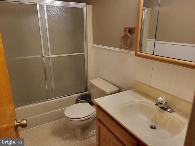 bathroom with vanity, wainscoting, a decorative wall, toilet, and bath / shower combo with glass door