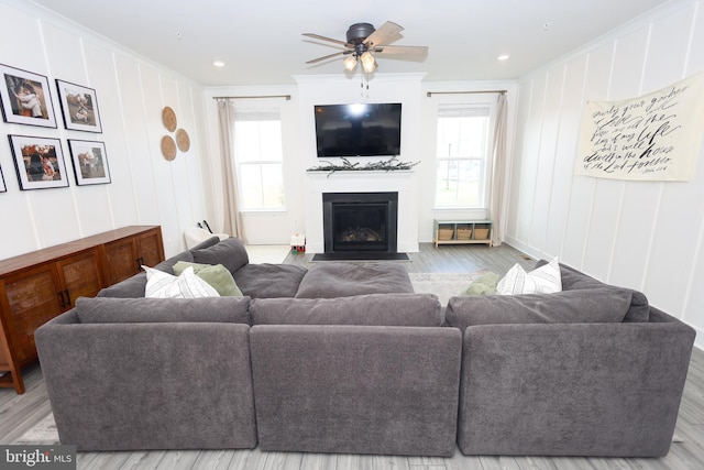 living area featuring a decorative wall, ceiling fan, wood finished floors, a fireplace with flush hearth, and crown molding