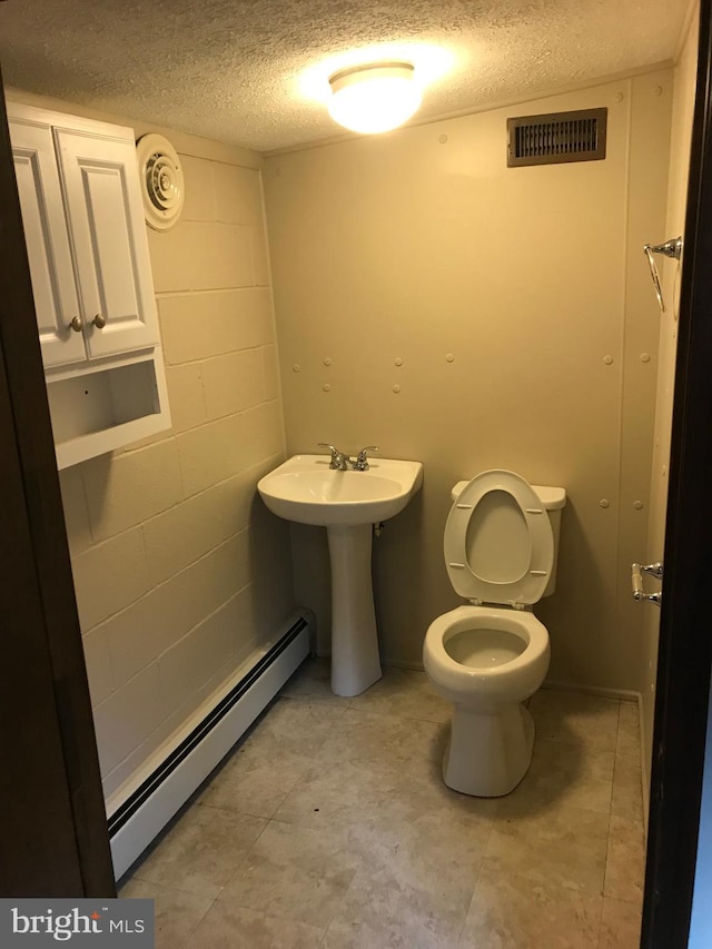 bathroom featuring visible vents, toilet, a textured ceiling, a baseboard radiator, and concrete block wall