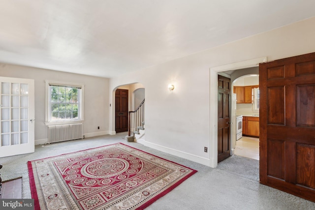 unfurnished living room featuring radiator heating unit, arched walkways, baseboards, and light colored carpet