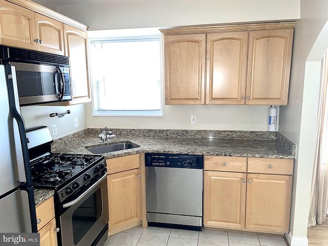 kitchen featuring light brown cabinetry, appliances with stainless steel finishes, a sink, dark stone countertops, and light tile patterned floors