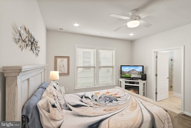 bedroom with ceiling fan, a lit fireplace, visible vents, wood finished floors, and baseboards