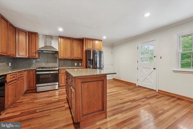 kitchen featuring wall chimney exhaust hood, stainless steel appliances, brown cabinetry, light wood-style flooring, and tasteful backsplash