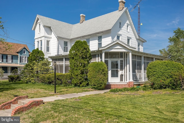 view of front of house featuring roof with shingles, a chimney, a sunroom, and a front yard