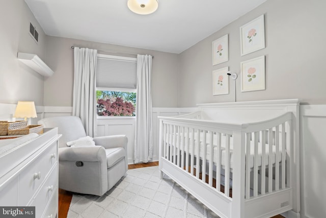 bedroom featuring visible vents, wainscoting, a decorative wall, a nursery area, and light wood finished floors