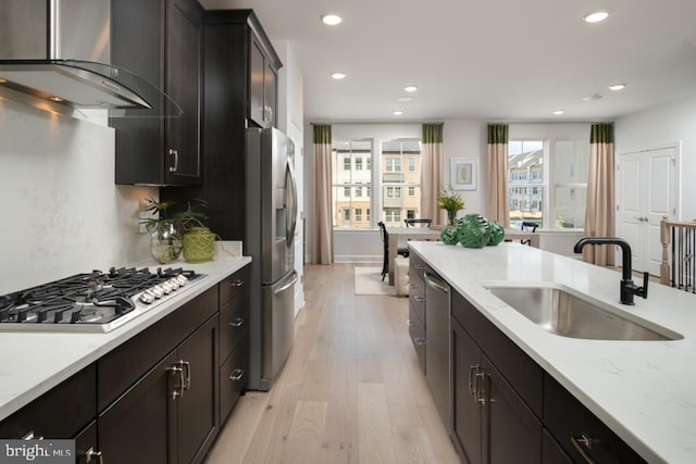 kitchen featuring appliances with stainless steel finishes, a sink, light wood finished floors, wall chimney exhaust hood, and recessed lighting