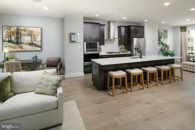 kitchen featuring wall chimney range hood, appliances with stainless steel finishes, a center island with sink, and open floor plan