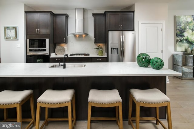 kitchen featuring dark brown cabinetry, a breakfast bar, wall chimney range hood, a sink, and stainless steel appliances