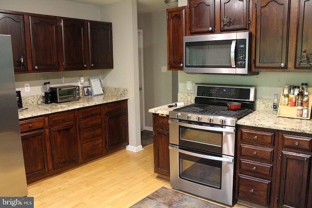 kitchen with dark brown cabinetry, light stone counters, stainless steel appliances, light wood-type flooring, and a toaster