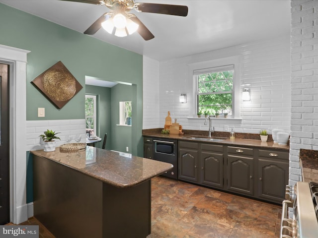 kitchen with a ceiling fan, stainless steel gas range oven, dark stone counters, a sink, and stone finish flooring