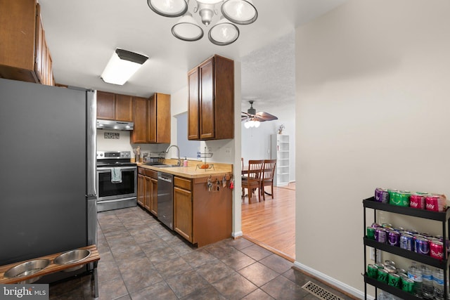 kitchen with brown cabinetry, light countertops, under cabinet range hood, a sink, and appliances with stainless steel finishes
