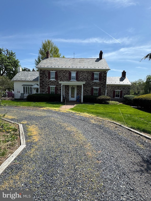 colonial-style house featuring a chimney, a front lawn, stone siding, and gravel driveway