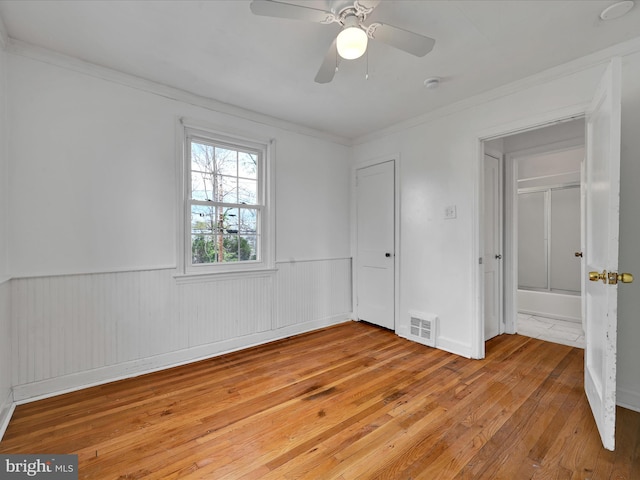 unfurnished bedroom featuring ceiling fan, visible vents, a wainscoted wall, light wood finished floors, and ornamental molding