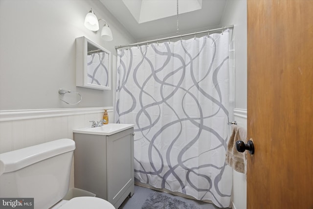 full bathroom with vanity, toilet, a shower with shower curtain, a wainscoted wall, and a skylight