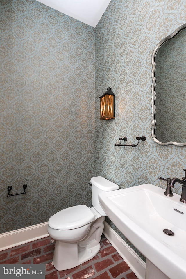bathroom with a sink, brick floor, toilet, baseboards, and wallpapered walls