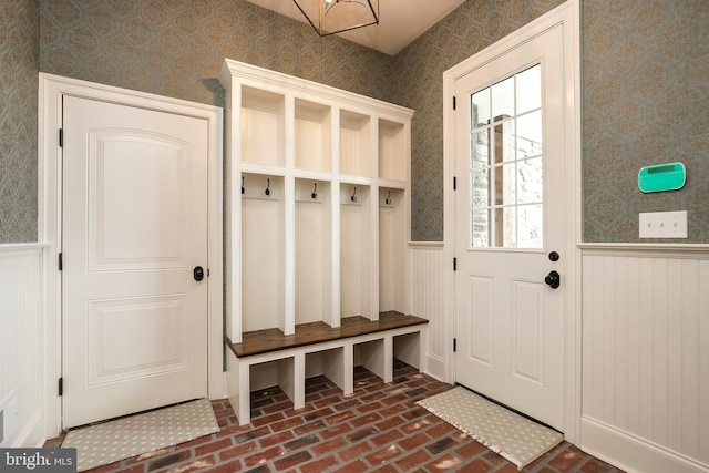 mudroom featuring an inviting chandelier, brick floor, wainscoting, and wallpapered walls
