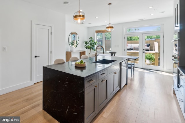 kitchen with a sink, an island with sink, french doors, light wood-style flooring, and dark stone countertops
