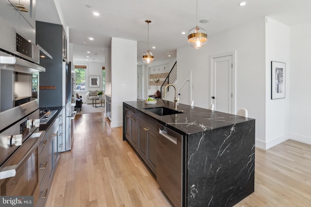 kitchen with appliances with stainless steel finishes, a sink, light wood finished floors, an island with sink, and dark stone counters