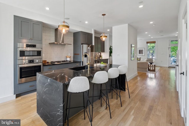kitchen with wall chimney range hood, appliances with stainless steel finishes, a sink, recessed lighting, and a center island with sink