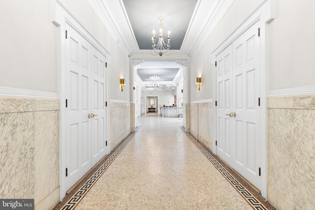 corridor featuring ornamental molding, wainscoting, tile walls, a tray ceiling, and a notable chandelier