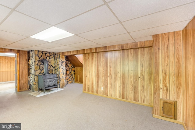finished below grade area featuring wooden walls, a drop ceiling, a wood stove, and carpet floors