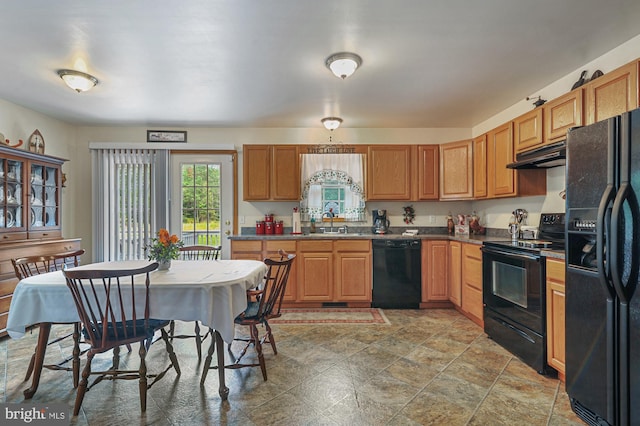 kitchen with stone finish floor, under cabinet range hood, brown cabinets, a sink, and black appliances