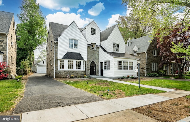 view of front of house with a front yard, an outdoor structure, a detached garage, a shingled roof, and stone siding