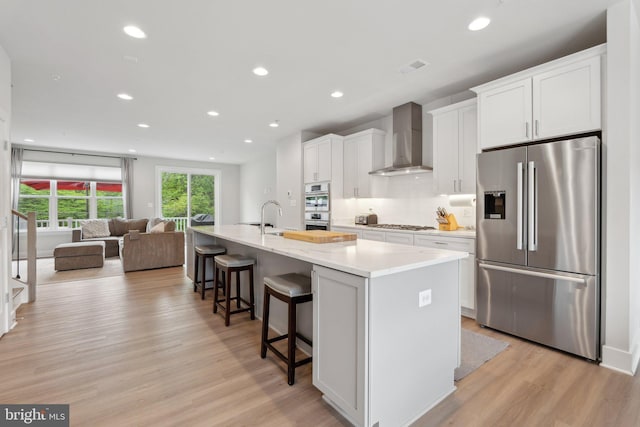 kitchen featuring a kitchen island with sink, a kitchen breakfast bar, wall chimney range hood, white cabinetry, and appliances with stainless steel finishes