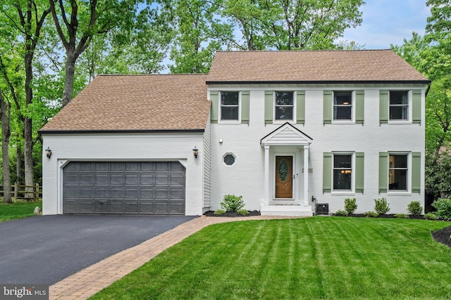 colonial inspired home with aphalt driveway, roof with shingles, brick siding, a front lawn, and an attached garage