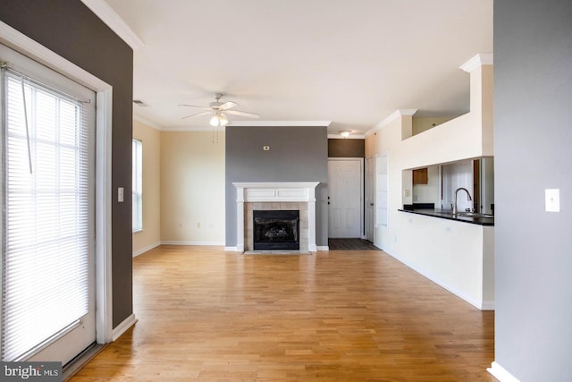unfurnished living room with ceiling fan, ornamental molding, a tiled fireplace, and light wood finished floors
