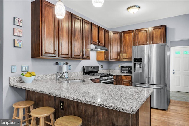 kitchen featuring a peninsula, light stone countertops, under cabinet range hood, a sink, and stainless steel appliances