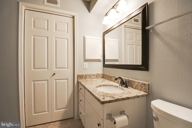 half bath featuring vanity, a textured wall, toilet, and visible vents