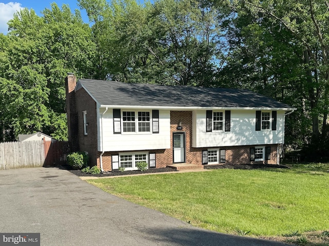 raised ranch featuring driveway, a chimney, brick siding, fence, and a front lawn