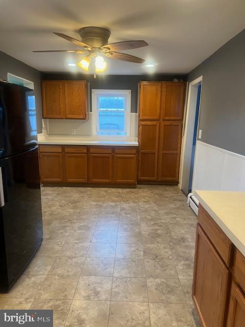 kitchen with a ceiling fan, freestanding refrigerator, brown cabinetry, wainscoting, and light countertops