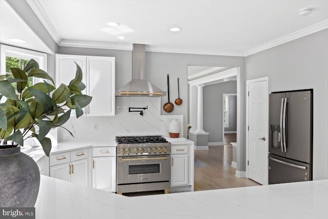 kitchen with appliances with stainless steel finishes, wall chimney range hood, ornate columns, healthy amount of natural light, and crown molding