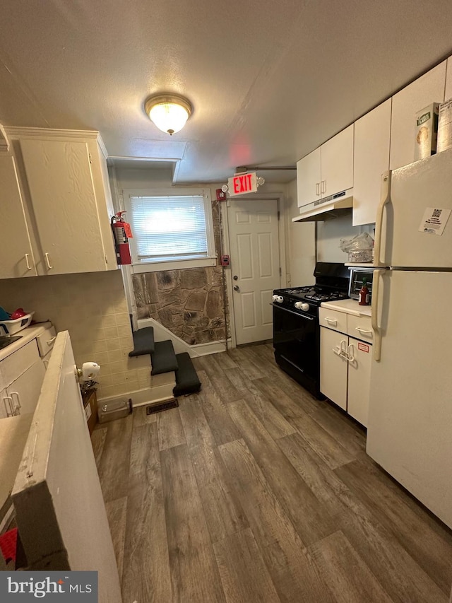 kitchen featuring wood finished floors, black gas range oven, freestanding refrigerator, under cabinet range hood, and white cabinetry