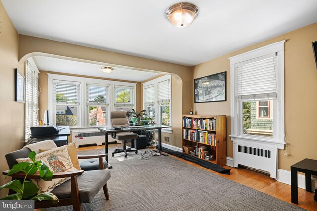 office area featuring arched walkways, baseboards, radiator heating unit, and wood finished floors