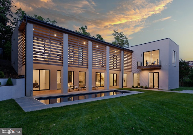 back of property at dusk with stucco siding, an outdoor pool, a balcony, a patio, and a yard