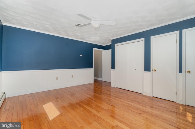 unfurnished bedroom with a ceiling fan, a baseboard radiator, a wainscoted wall, wood finished floors, and multiple closets